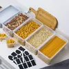 A collection of clear plastic food storage containers filled with various dry goods like pasta, cereal, and oats, arranged on a white countertop next to a wooden cutting board and a set of chalkboard labels. Produktbild-Vorschau