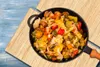 A close-up overhead shot of a pan filled with mixed vegetables, placed on a wooden cutting board which is on a blue wooden surface. Náhled obrázku produktu