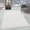 A cream-colored rug is placed on a light gray wooden floor in a living room setting. A grey armchair is visible to the right, and a framed picture of the Eiffel Tower is on the wall to the left. A large window shows a snowy outdoor scene. Produktbild-Vorschau