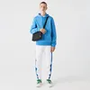 A man wearing a blue hoodie and white pants with blue stripes, carrying a black bag, stands against a white background. Voir les images du produit
