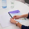 A person's hands are shown placing a document into a binder on a white table. There is a bottle of water and a glass of water in the background. Náhled obrázku produktu