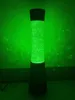 A tall, cylindrical lava lamp with green liquid and bubbles, lit from within, casting a green glow on the wall behind it. Anteprima dell’immagine prodotto