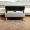 A grey storage bench with its lid open, revealing two white pillows inside, placed on a wooden floor in front of a bed. Náhled obrázku produktu