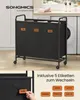 A laundry basket with three compartments and wheels, placed in a laundry room setting with a washing machine in the background. The image also shows labels for the compartments and a separate display of interchangeable labels. Produktbild-Vorschau