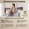 A woman is sitting on a bed wrapped in a gray heated blanket, holding the controller. Produktbild-Vorschau