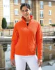 A woman wearing an orange fleece jacket and white pants stands outdoors near a body of water with buildings in the background. Produktbild-Vorschau