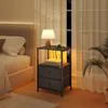 A bedside table with a lamp, books, and charging cables next to a bed. Produktbild-Vorschau