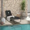 A black foldable lounge chair with a headrest and a towel is placed on a wooden deck next to a swimming pool. A stone wall serves as the background, with a potted plant and a small stool with books. Produktbild-Vorschau