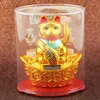 A golden Maneki-neko (lucky cat) figurine with a solar-powered waving paw, displayed in a clear plastic case on a red base, set against a textured brown background. Produktbild-Vorschau