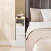A narrow white nightstand with a wooden top, placed next to a bed with brown headboard and beige and white bedding. Voir les images du produit