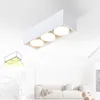 A white rectangular ceiling light with three circular light sources is shown against a white ceiling. A faint sketch of the light and its illumination pattern is in the lower left corner. The background shows a blurred interior scene with a sofa and furnit Produktbild-Vorschau