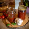 Two glass jars filled with red sauce, a head of garlic, and basil leaves are arranged on a wooden board, with tomatoes in the background. Produktbild-Vorschau