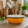 A bright orange enamel bowl sits on a countertop in a kitchen setting, with other kitchen items like copper pots and pasta in the background. Podgląd zdjęcia produktu