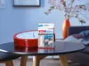 A tesa Powerstrips adhesive strip package is placed on a dark round table next to a red clock. In the background, a vase with flowers and a sofa are visible. Anteprima dell’immagine prodotto