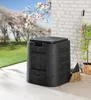 A black compost bin sits on a patio next to a brick wall, with a broom leaning against the wall and a potted plant nearby. A grassy lawn and trees with white blossoms are in the background. Produktbild-Vorschau