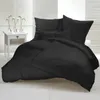 A neatly made bed with black bedding, including pillows and a duvet, is shown in a bright room. A small white nightstand with three vases of white flowers is to the left of the bed. Produktbild-Vorschau