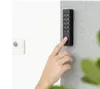 A hand is shown pressing a button on a black Nuki Keypad 2.0 mounted on a textured gray wall. Produktbild-Vorschau