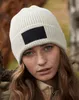 A close-up shot of a young woman wearing a cream-colored beanie with a black patch, outdoors in a natural setting. Anteprima dell’immagine prodotto