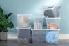 Several clear plastic storage boxes are stacked and arranged in a room with a blue wall and a plant on the left. Anteprima dell’immagine prodotto