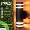 A black outdoor wall light with two illuminated bands is mounted on a wooden wall, with a green hedge in the background. Rain is falling. Anteprima dell’immagine prodotto