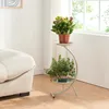 A two-tiered plant stand with two potted plants is placed next to a sofa in a well-lit room with a window. Podgląd zdjęcia produktu