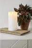 A white LED candle sits on a wooden tray next to a potted plant. Produktbild-Vorschau