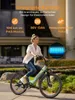 A woman is riding a mint green electric bicycle in an outdoor setting with a building in the background. Produktbild-Vorschau