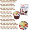 A collection of small jars with fruit-themed lids, a recipe book, and a decorative plate, all arranged on a white background. Podgląd zdjęcia produktu