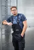 A man wearing overalls and a t-shirt leans against a metal structure. Náhled obrázku produktu