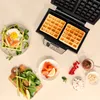 A waffle maker with two waffles, a prepared waffle with toppings, and various ingredients for waffles are laid out on a table. Anteprima dell’immagine prodotto