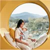 A woman is sitting in a circular window seat with a baby in a carrier, looking out at a mountainous landscape. Produktbild-Vorschau