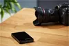 A black external hard drive and a DSLR camera are placed on a wooden table. Produktbild-Vorschau
