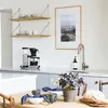 A kitchen counter with a coffee maker and shelves above, and a dining table set for a meal in the foreground. Náhľad obrázku produktu