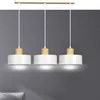 A three-light pendant lamp with white cylindrical shades and natural wood accents, suspended from a light wood bar against a gray wall. Náhľad obrázku produktu