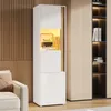 A tall, narrow white cabinet with wood accents and internal LED lighting, displaying books and decorative items. It is placed in a modern living room setting next to a sofa and a larger wooden shelving unit. Anteprima dell’immagine prodotto