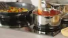 A close-up shot of a small saucepan with red sauce being stirred with a wooden spoon, placed on a black induction cooktop. A larger frying pan with food is visible in the background. Anteprima dell’immagine prodotto