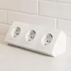 A white triple electrical socket outlet is placed on a white countertop in front of a white tiled wall. Produktbild-Vorschau