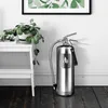 A silver fire extinguisher stands on a dark wooden floor next to a white shelf with a plant on it. A framed artwork with green leaves is on the wall above. Produktbild-Vorschau