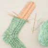 A knitting ruler is placed on a partially knitted sock, with a ball of yarn and knitting needles nearby. Produktbild-Vorschau