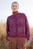A woman stands outdoors in tall grass, wearing a striped cardigan and brown pants. Produktbild-Vorschau