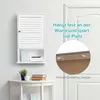 A white bathroom cabinet with an open shelf is mounted on a white wall above a white console table. The cabinet has a slatted door and a small knob. The open shelf contains a framed picture and some folded items. The console table has a potted plant, an ap Náhled obrázku produktu