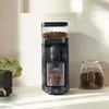 A black coffee grinder with coffee beans in the hopper and ground coffee in the collection bin, placed on a kitchen counter next to a potted plant and a jar of coffee beans. Produktbild-Vorschau