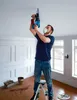 A man is using a drill on the ceiling while standing on a step ladder. Produktbild-Vorschau
