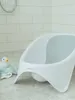 A white baby bathtub is shown in a bathroom setting with tiled walls and floor. A rubber duck and towels are placed next to the bathtub. Produktbild-Vorschau