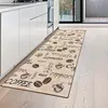 A kitchen runner rug with coffee-themed patterns is placed on a light wooden floor next to white kitchen cabinets. Voir les images du produit
