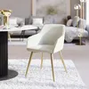 A cream-colored chair with gold legs is placed on a white rug in a living room setting. A round table with a vase and a cup is visible in the foreground, and a sofa and floor lamp are in the background. Produktbild-Vorschau