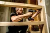 A man is using a cordless drill to screw a piece of wood into a wooden frame. Anteprima dell’immagine prodotto