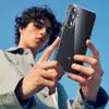A young man is holding a clear phone case with a Samsung phone inside, against a bright blue sky. Produktbild-Vorschau