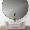 A close-up shot of a transparent glass vessel sink with a gold faucet, set against a round mirror and a wooden surface. Anteprima dell’immagine prodotto