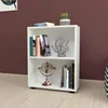 A white bookshelf with two shelves, filled with books and decorative items, placed on a light-colored wooden floor against a dark red wall. Anteprima dell’immagine prodotto